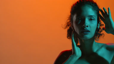 Curly-haired latina with teal light touches face on orange bg