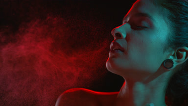 Latin beauty with red and cyan light sprays red mist on black bg