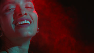 Smiling latin woman in cyan-red light sprays red mist on black