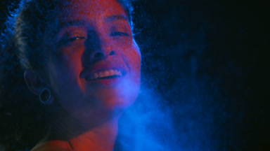 Cheerful latin woman in red-blue light sprays blue mist on black