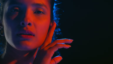 Latin beauty in red-blue light strokes jawline on black backdrop