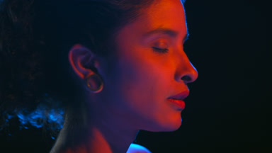 Brown-skinned woman in red-blue light strokes skin on black bg