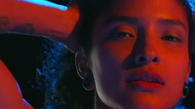 Brown-eyed woman in red-blue light holds hair looks at camera
