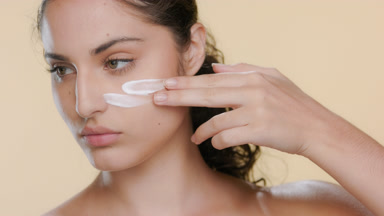 Fair-skinned model applies cream on cheek, looks aside